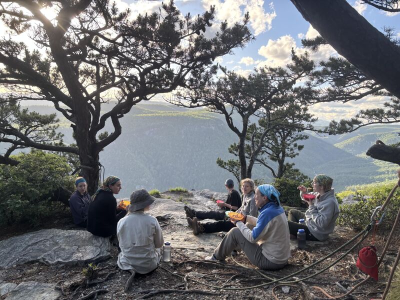A group of people are sitting on a rocky outcrop, possibly a mountain peak, enjoying a meal with a scenic view. They are surrounded by trees, and in the distance, there are mountains and valleys. The sky is partly cloudy, and the overall atmosphere suggests they are having a picnic or break during a hike.